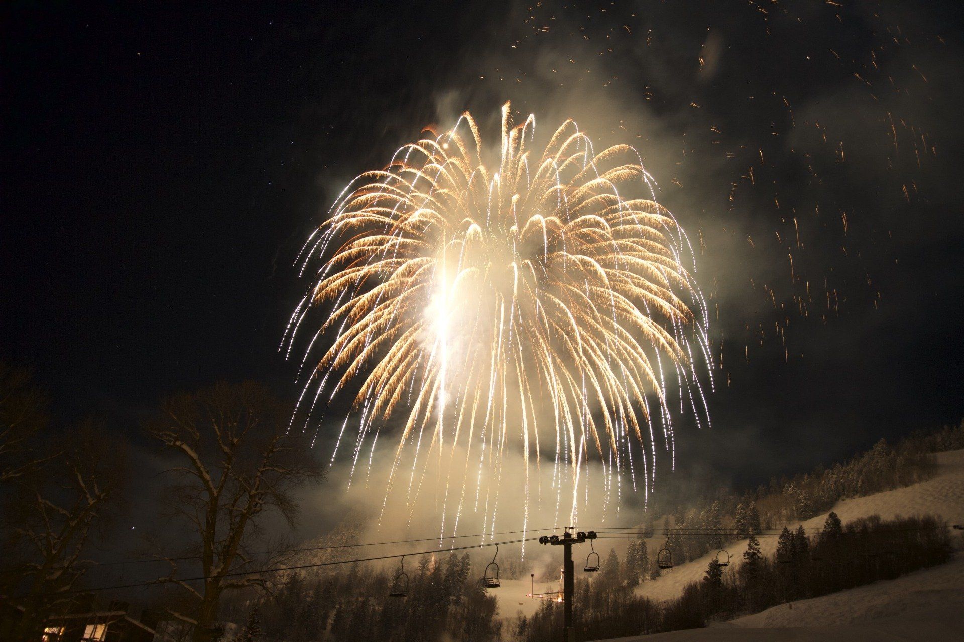 Fireworks bursting in the night sky over a snow-covered mountain. The display is bright yellow and white, illuminating the scene.