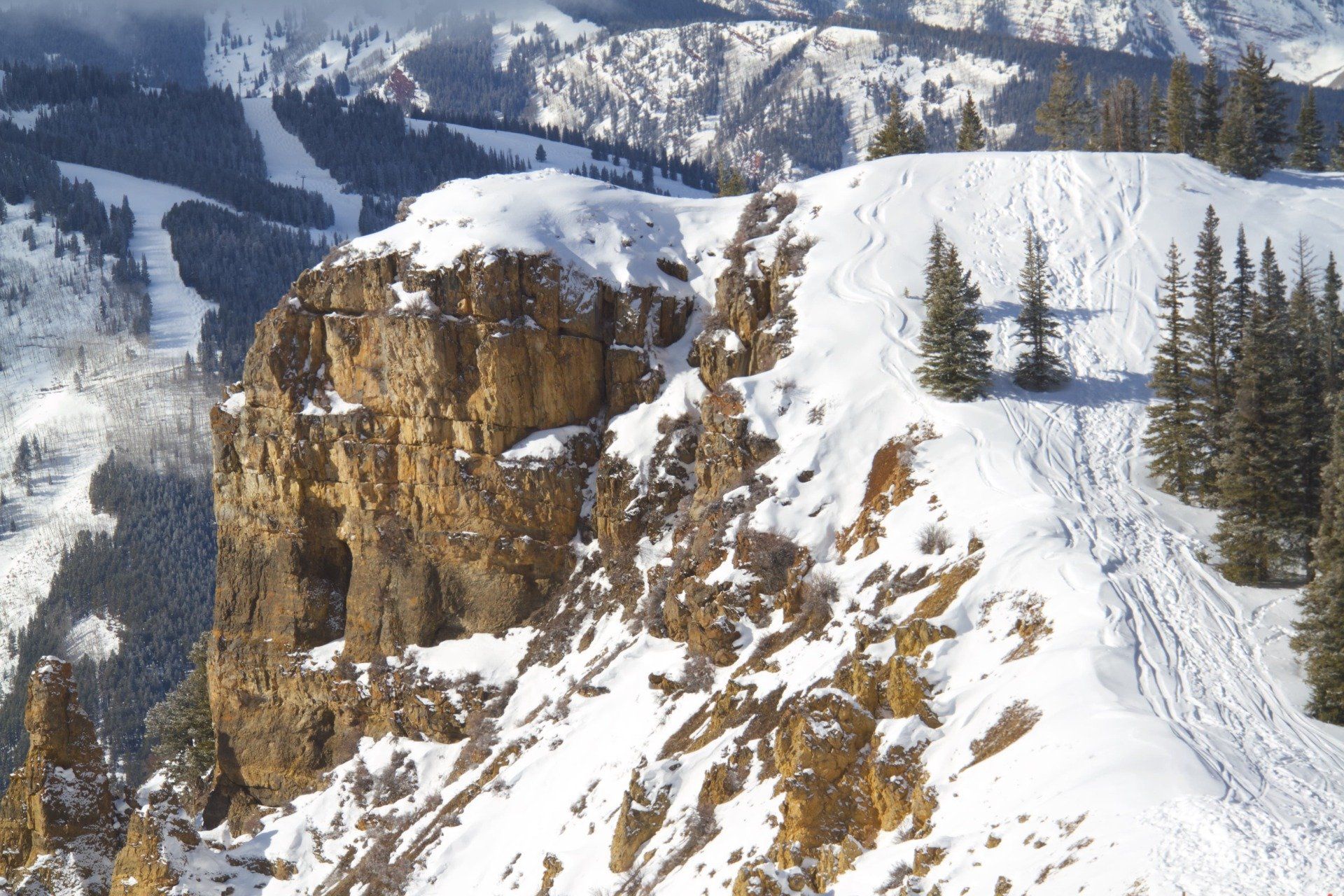 Snow-covered mountain cliff with a ski slope visible in the background. Sunlight illuminates the rock face and snow.