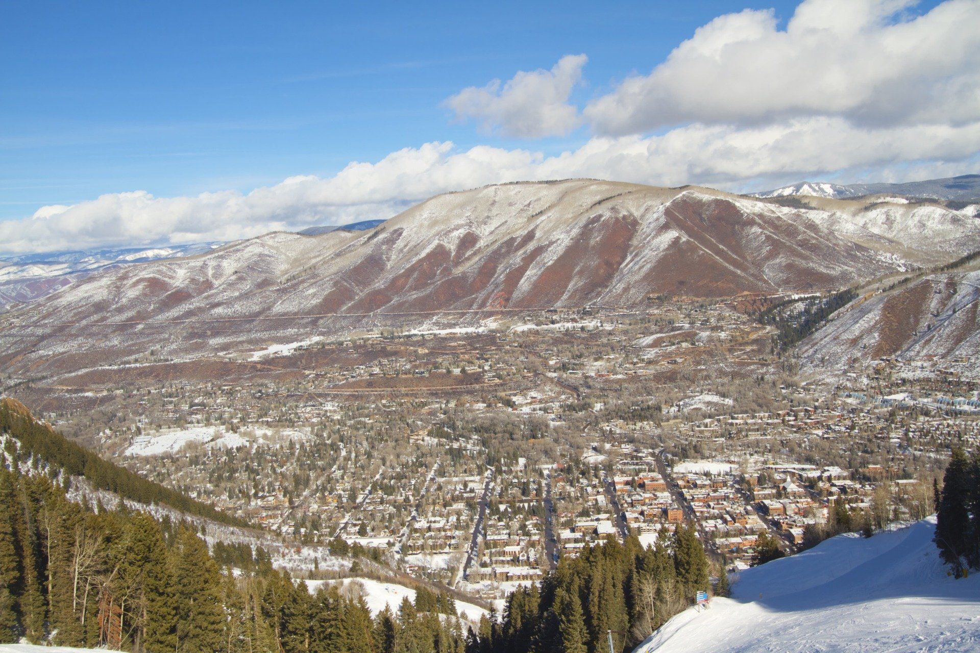 Snowy mountain landscape overlooking a town nestled in a valley, under a blue sky with scattered clouds.