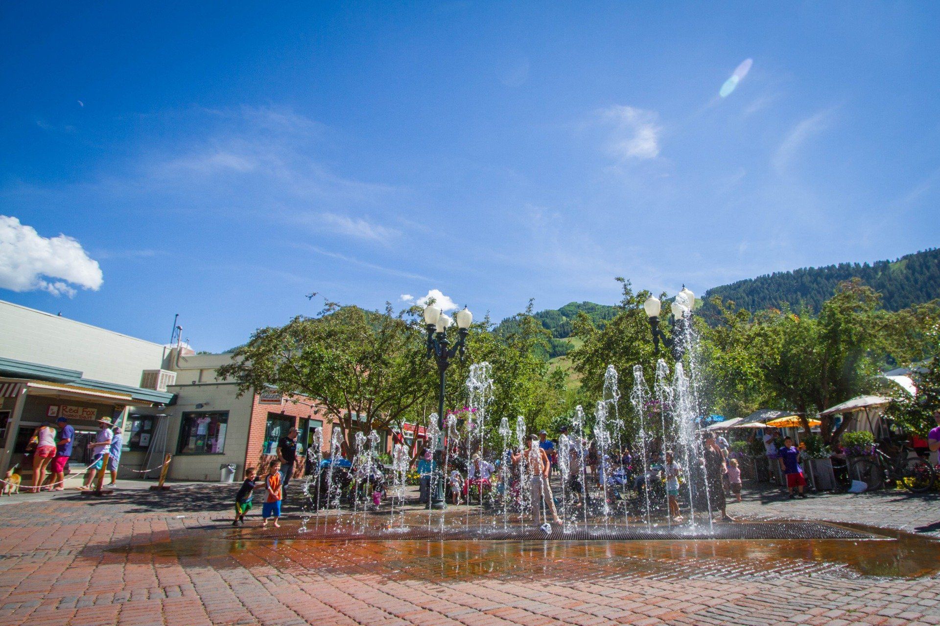 A sunny day at a town square with a water fountain, people, and shops. Trees and mountains visible in the background.
