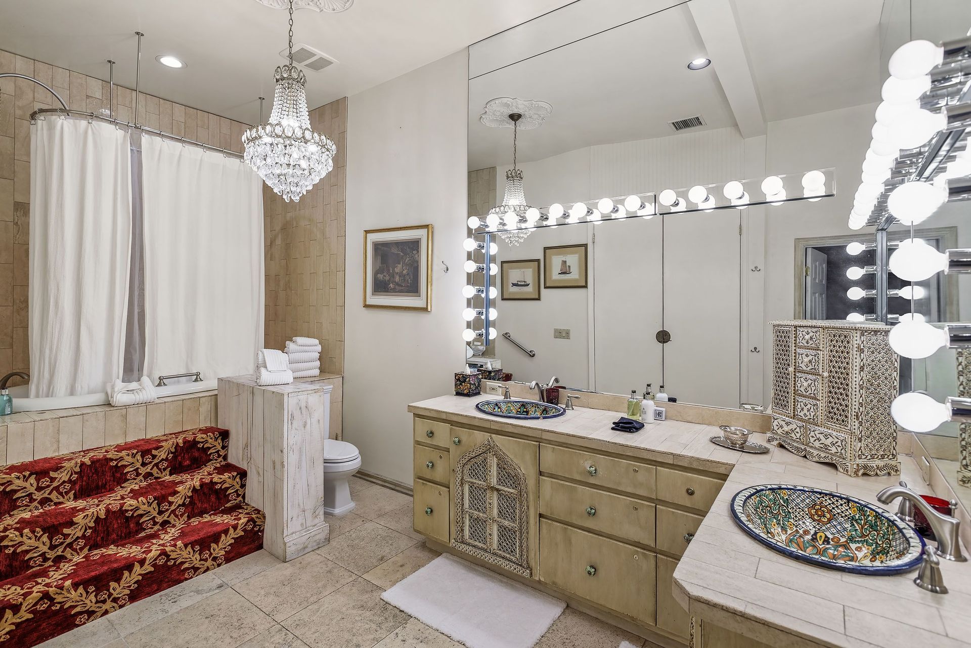 Luxurious bathroom with a red-carpeted tub, ornate vanity, and crystal chandeliers; cream walls and marble floor.