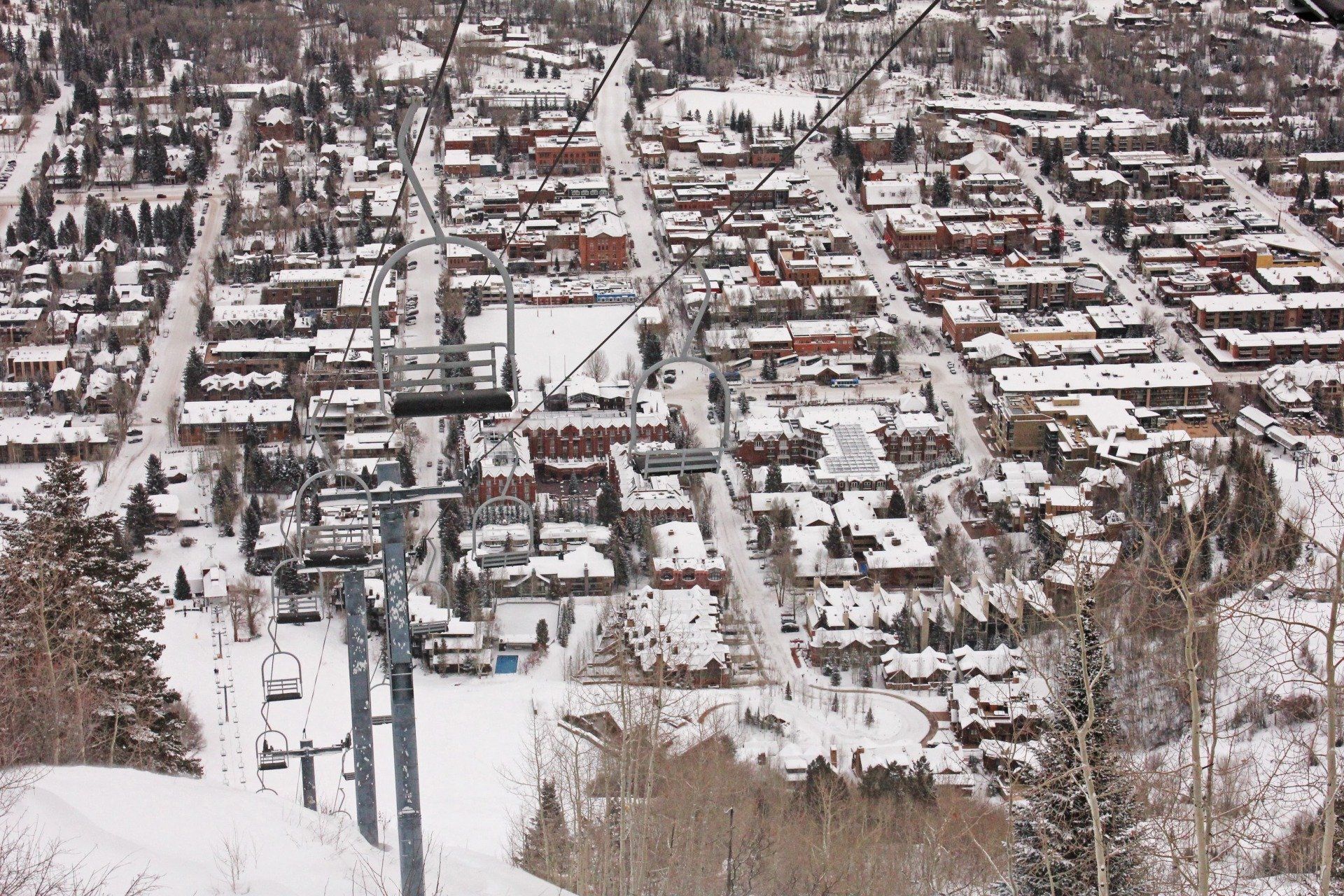 Overhead view of a snow-covered Aspen with a ski lift in the foreground. Buildings are brown and white, with scattered evergreen trees.