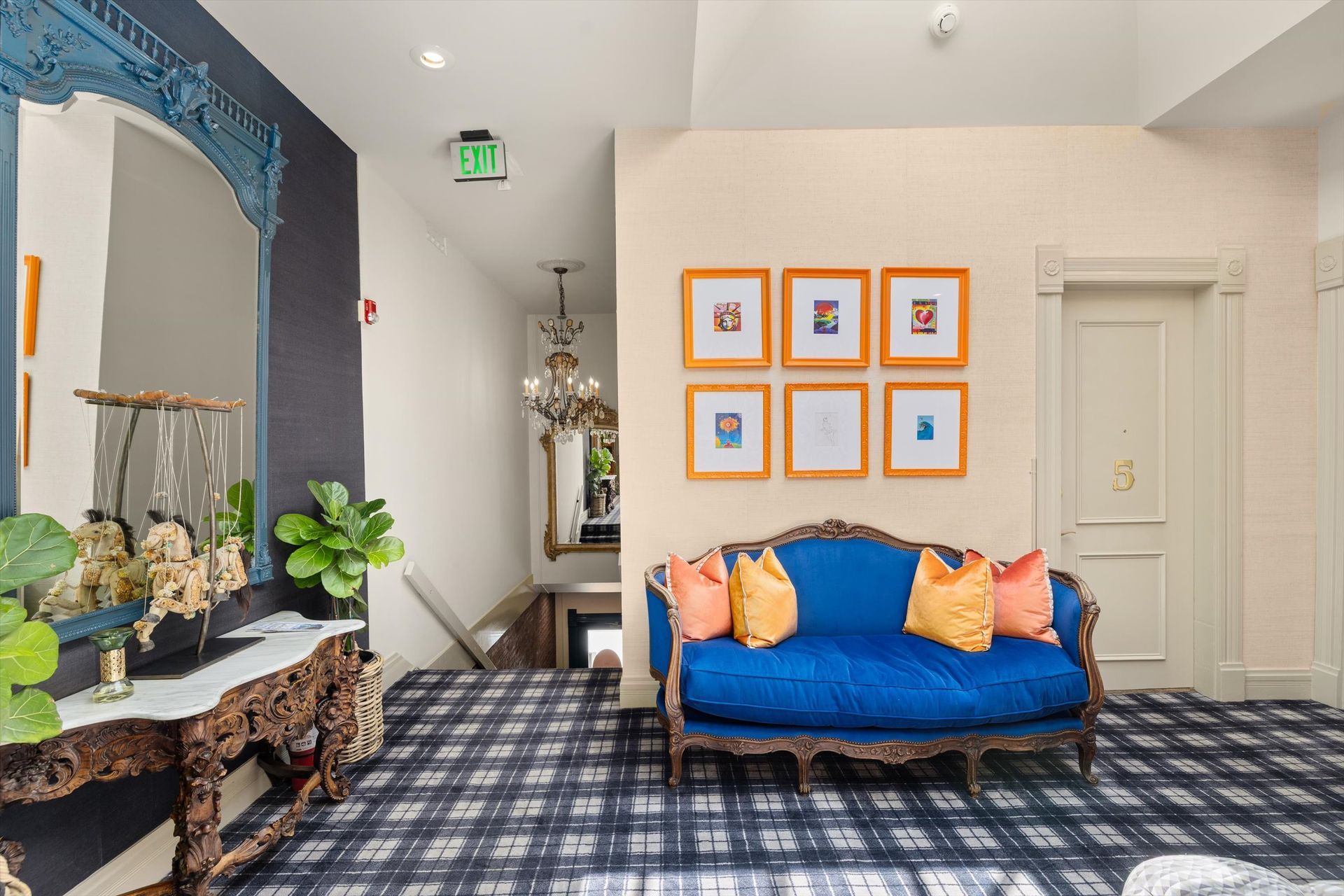 A blue velvet couch with orange pillows sits on a plaid carpet. A hall with a mirror, art, and an exit sign is behind it.