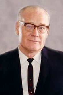 Portrait of an older man wearing glasses and a dark suit. He is smiling slightly.