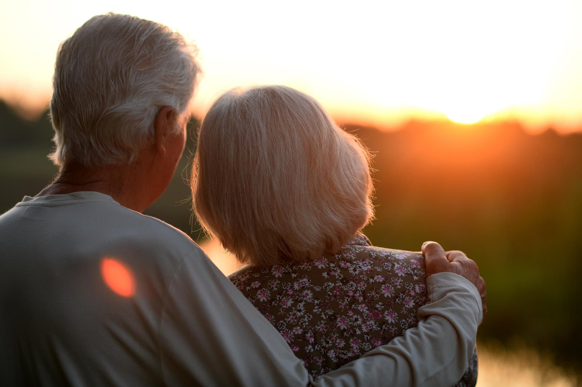 Elderly couple embracing, watching the sunset. Man's arm around woman's shoulder. Golden light, natural setting.
