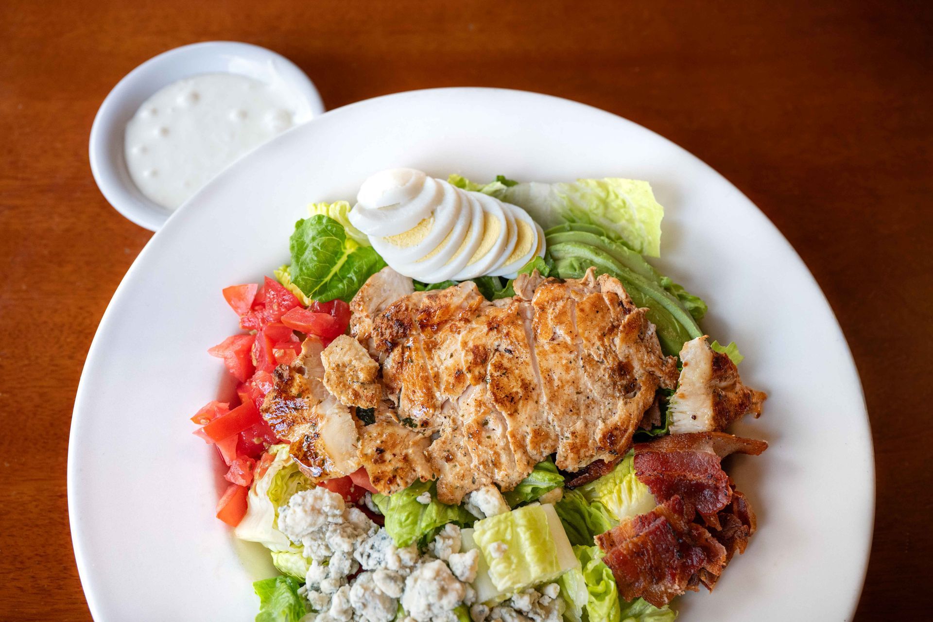 Plate of salad with grilled chicken, avocado slices, tomatoes, and a side of dressing on a white table