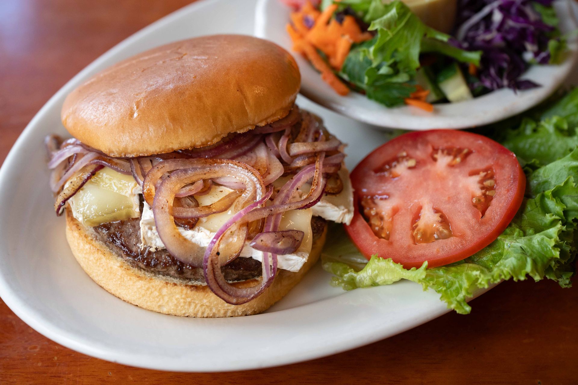 Burger with caramelized onions, melted cheese, tomato, and lettuce on a white plate, with salad in the background