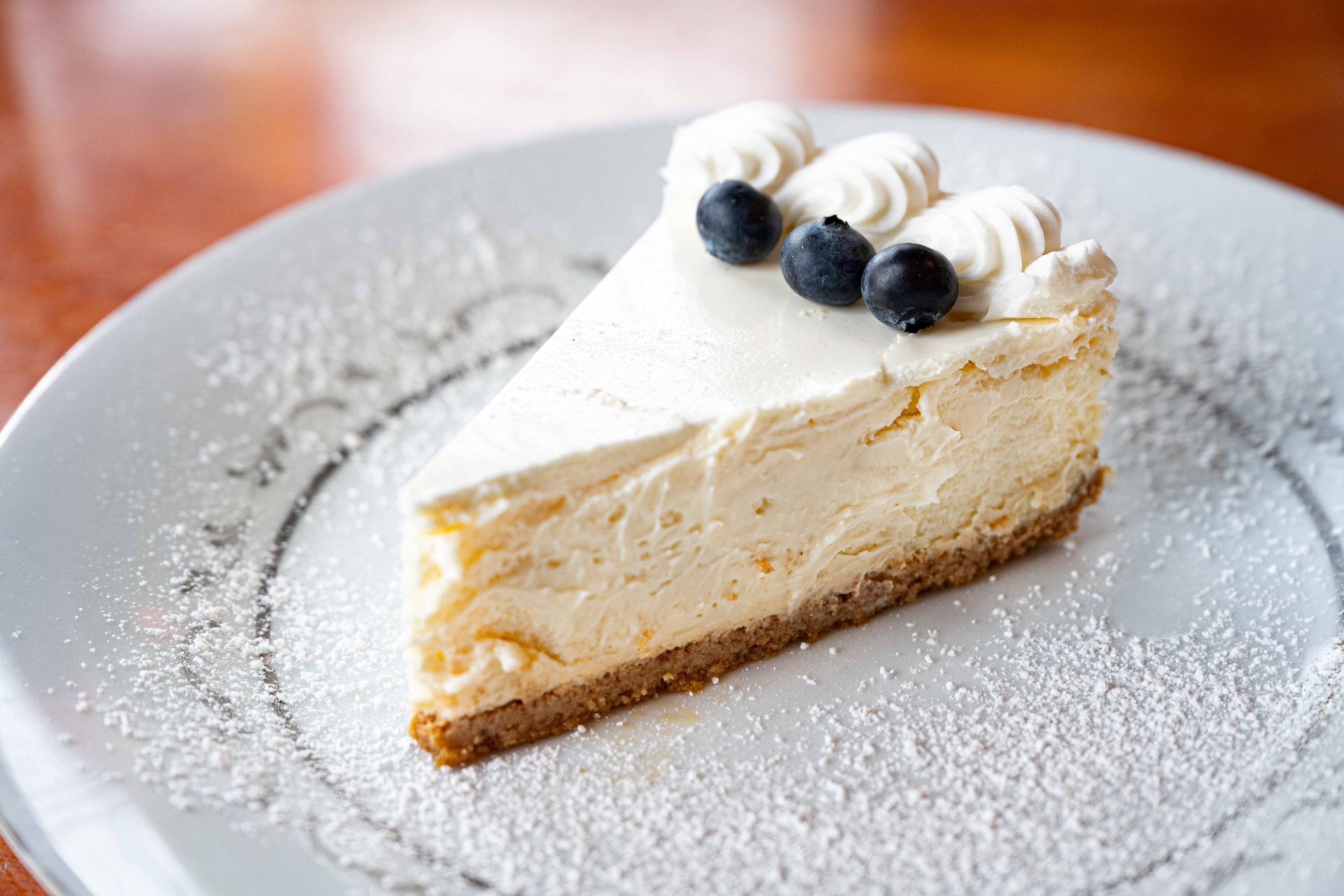 Slice of cheesecake on a white plate, topped with blueberries and whipped cream, dusted with powdered sugar
