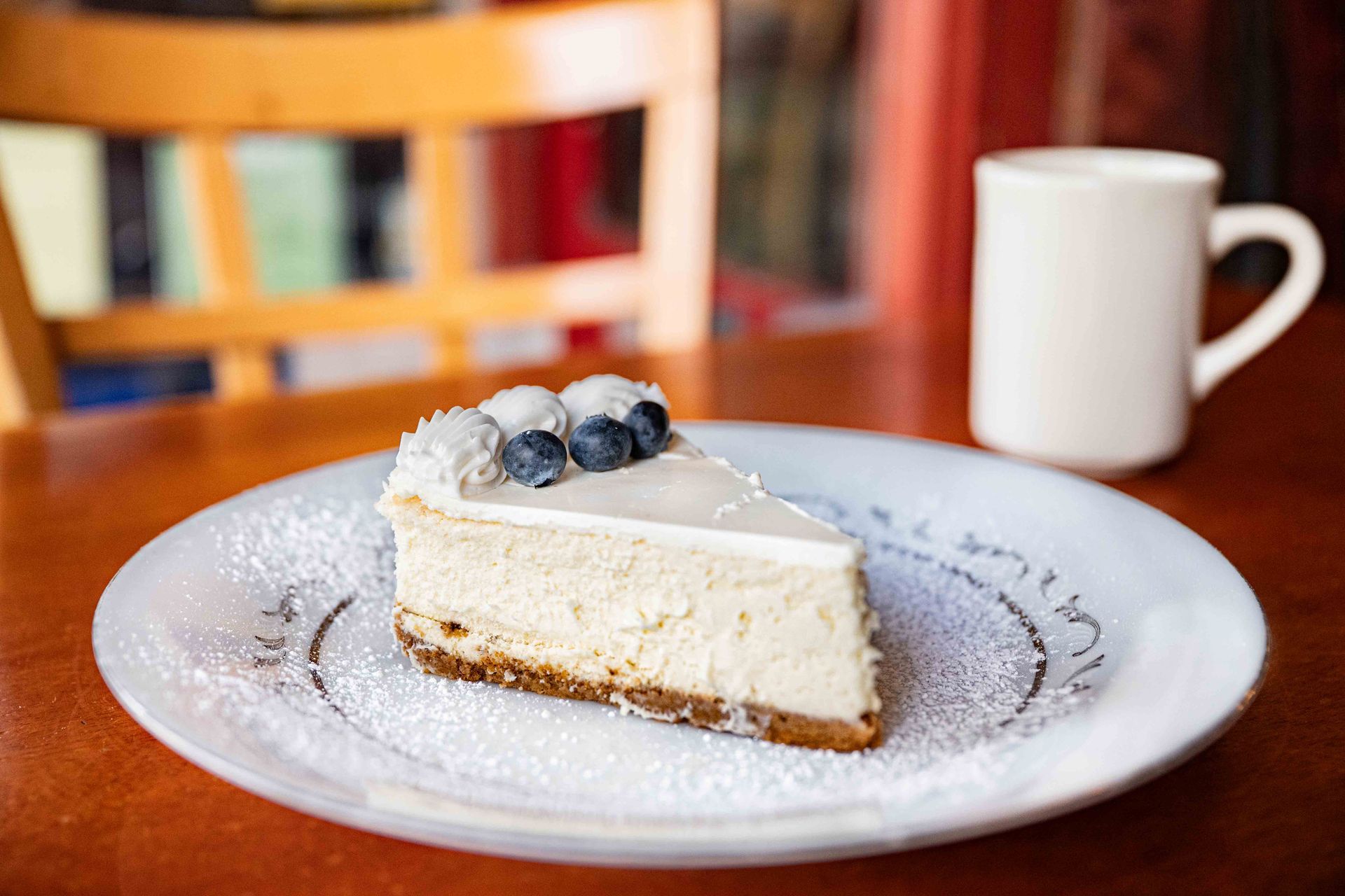 Slice of cheesecake topped with blueberries on a white plate, with a coffee mug in the background.