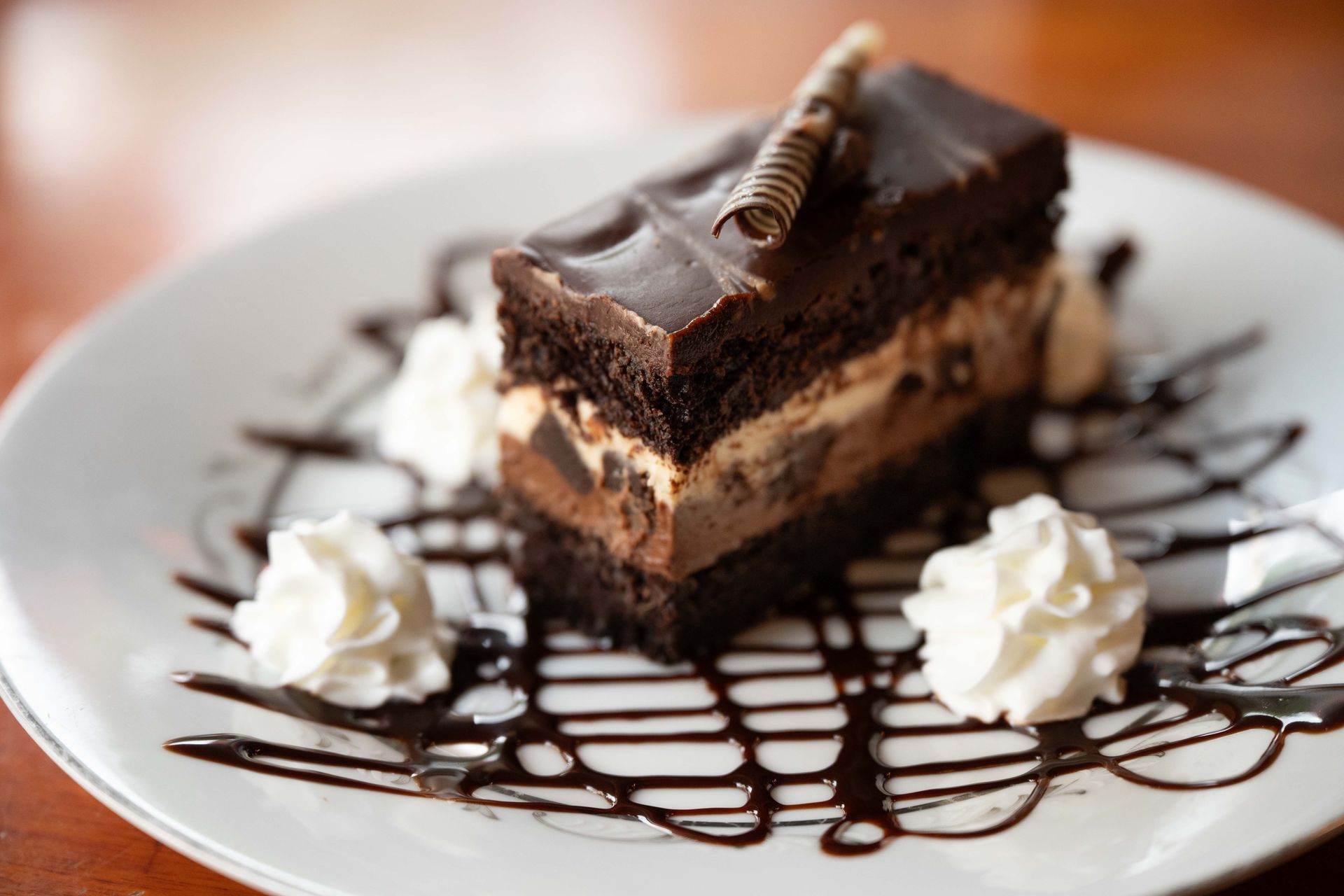 Chocolate cake slice on a white plate, drizzled with chocolate sauce and topped with whipped cream swirls