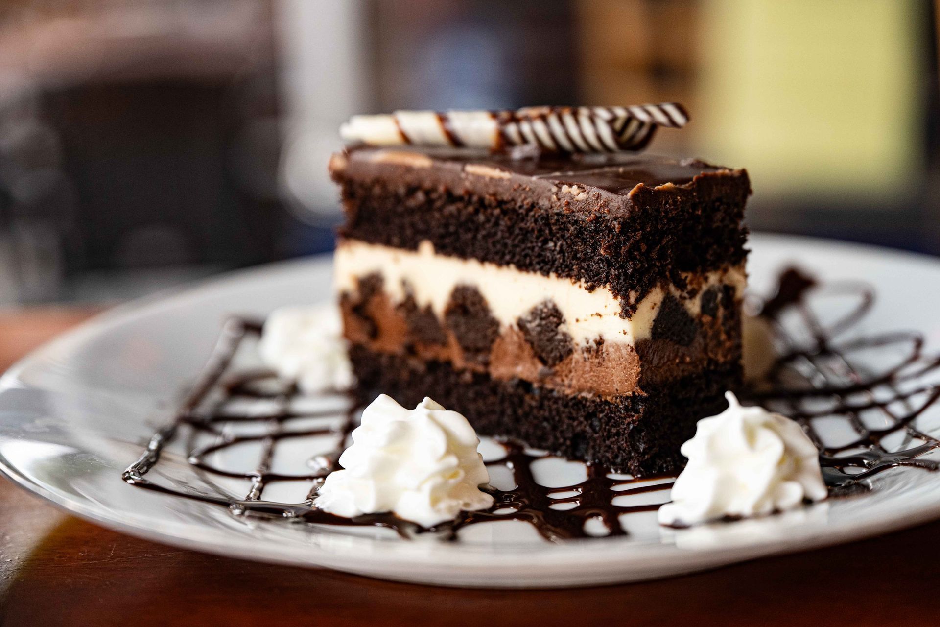Chocolate cake slice with cream layers and whipped cream, drizzled with chocolate sauce on a plate