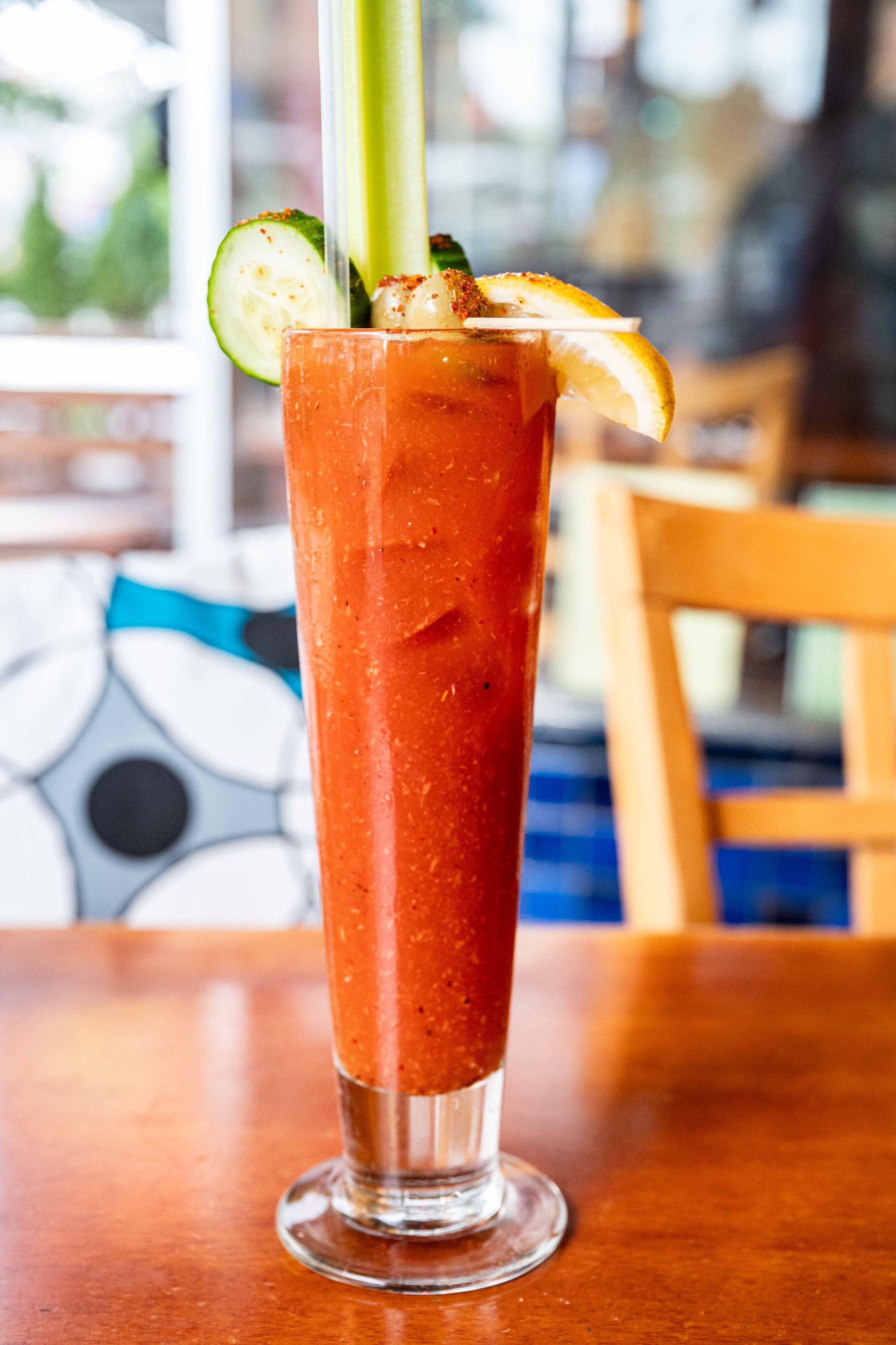 Tall red cocktail in a glass with cucumber and citrus garnish on a wooden table, in a café setting