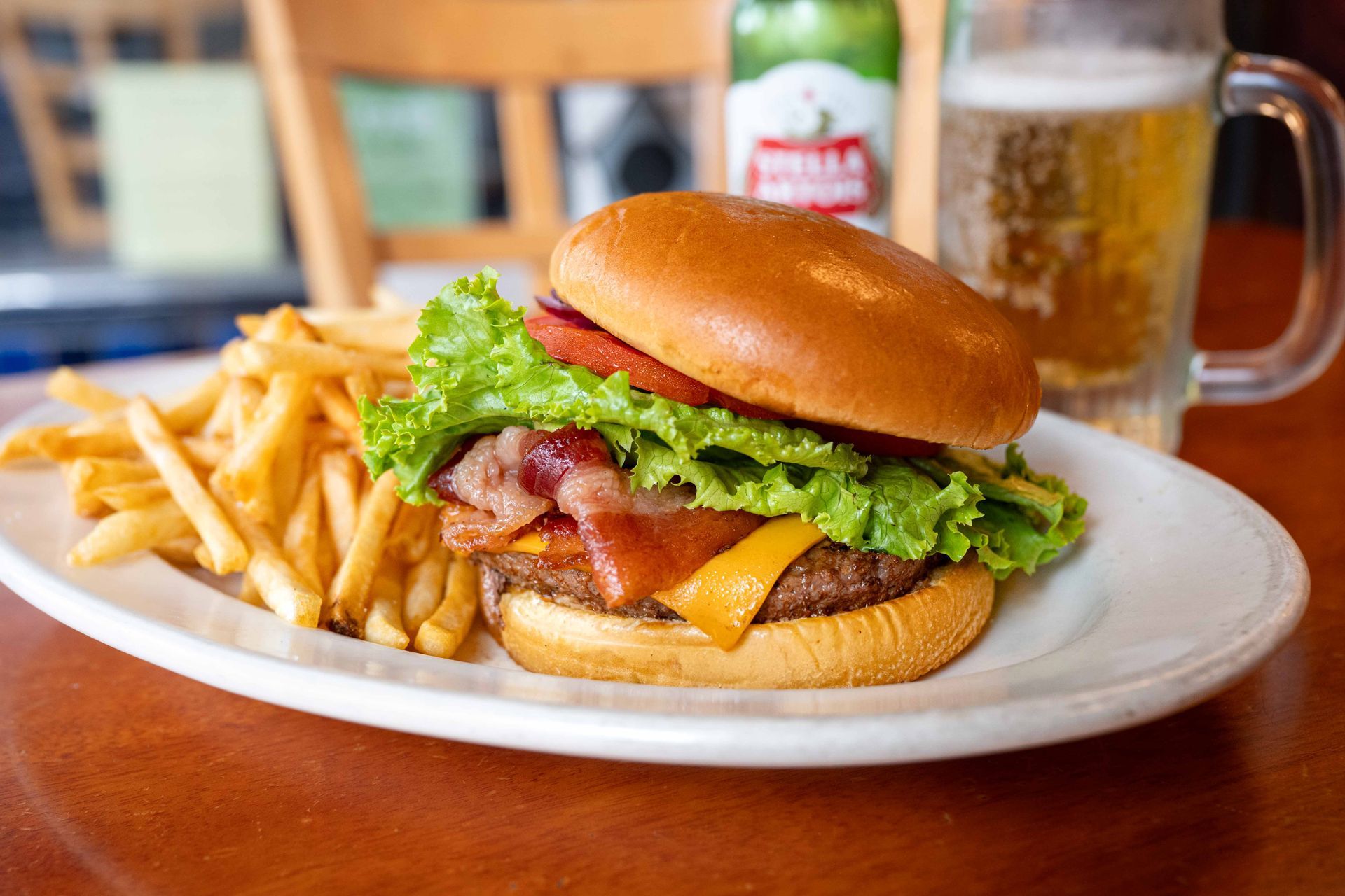 Burger with lettuce, bacon, cheese, and fries on a plate, with a beer mug in the background.