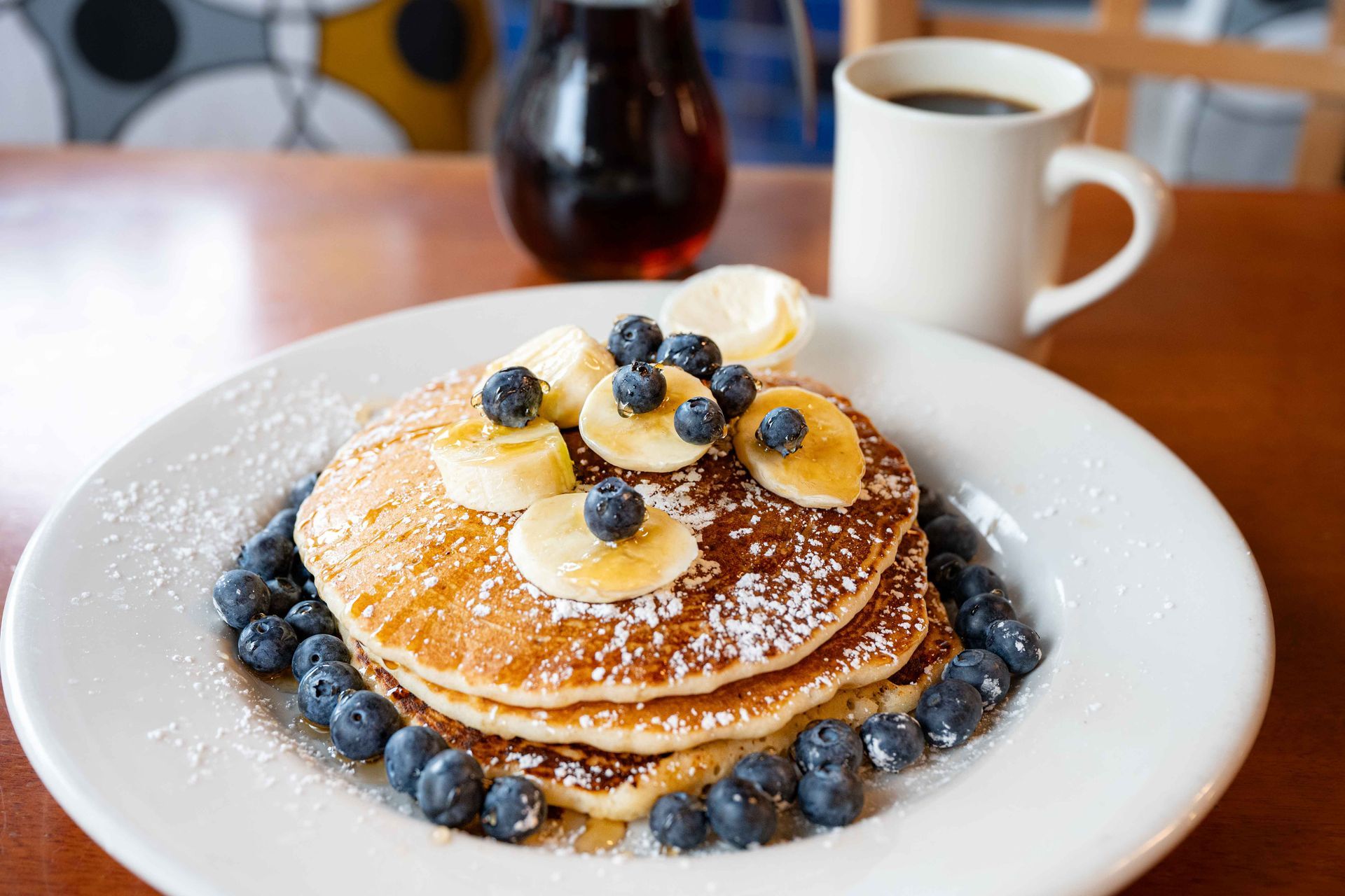 Blueberry pancakes dusted with powdered sugar on a white plate, with coffee in the background