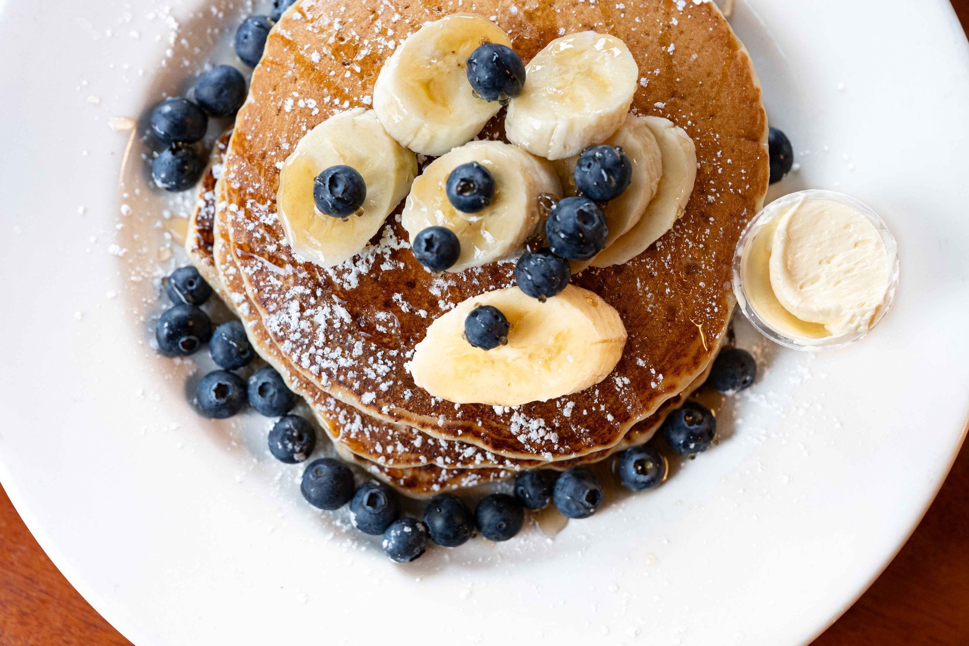 Stack of pancakes topped with banana slices, blueberries, and powdered sugar on a white plate