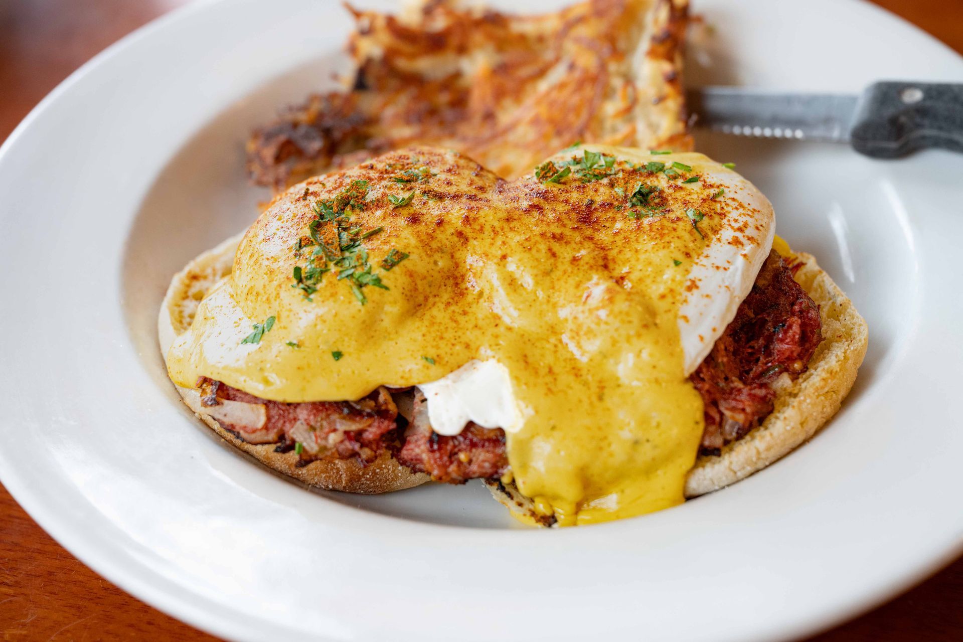 Eggs Benedict with hollandaise sauce on a white plate, garnished with herbs, alongside breakfast potatoes.