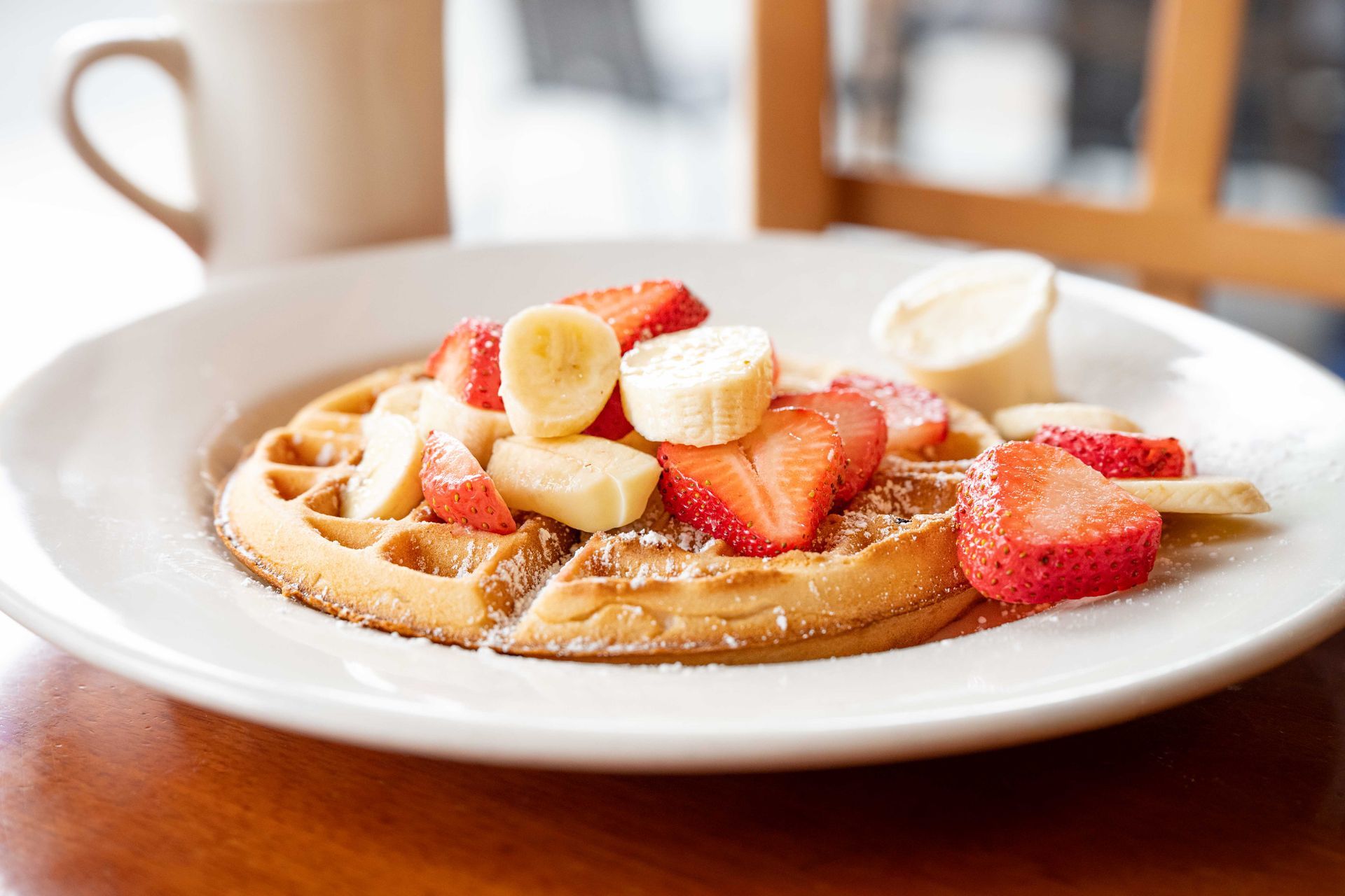 Waffle topped with sliced strawberries and bananas on a white plate on a wooden table