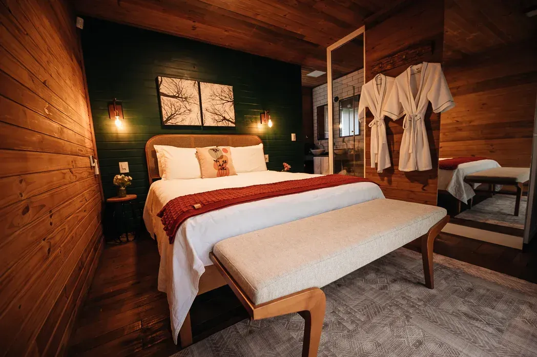 Um quarto com cama, banco, espelho e roupões pendurados na parede.
