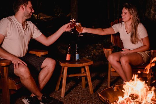 Um homem e uma mulher estão sentados ao lado de uma fogueira brindando com cerveja.