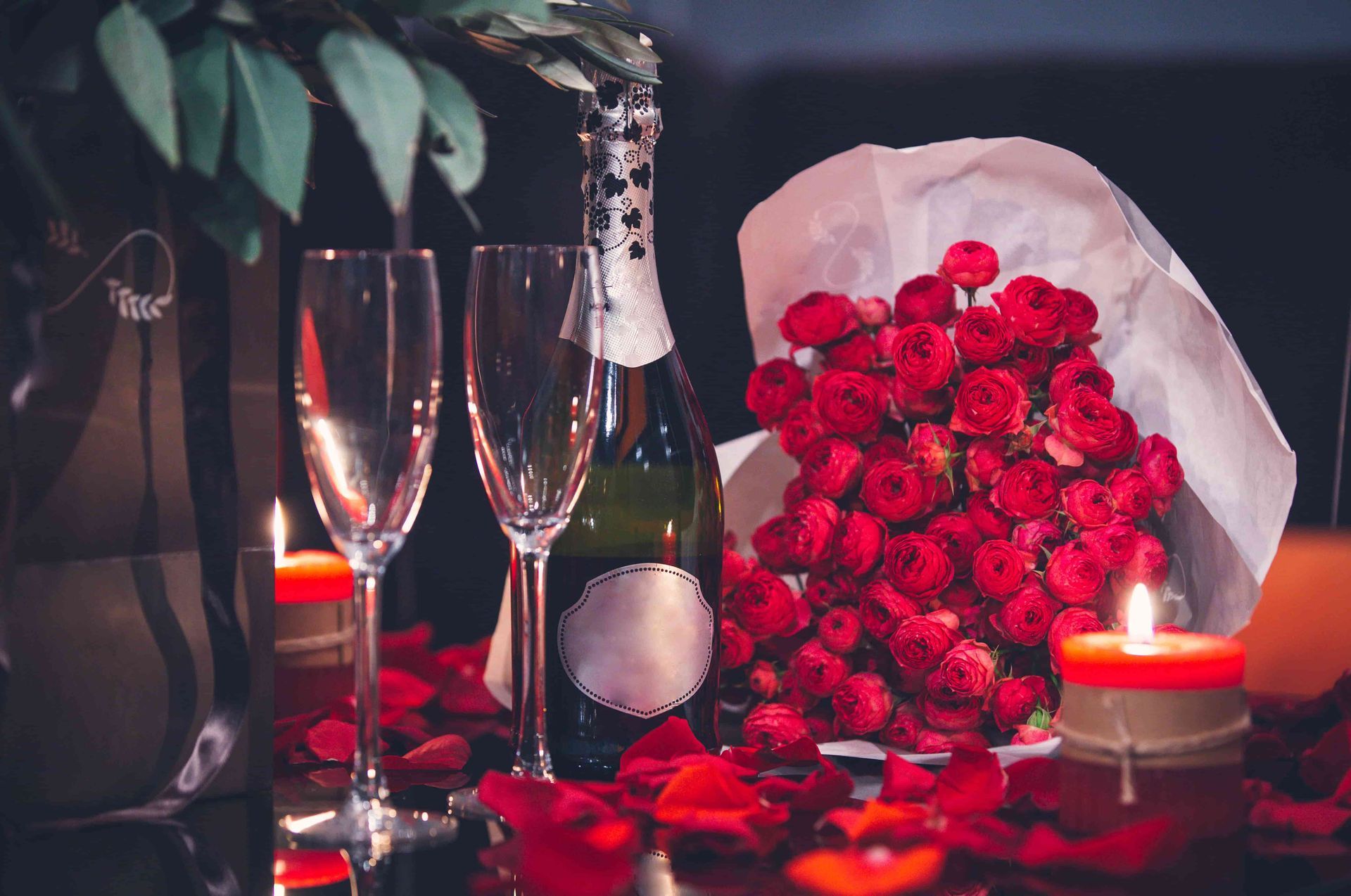 Uma garrafa de champanhe, duas taças, um buquê de rosas vermelhas e velas sobre uma mesa.