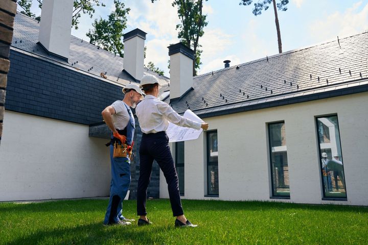 A man and a woman are standing in front of a house looking at a blueprint.