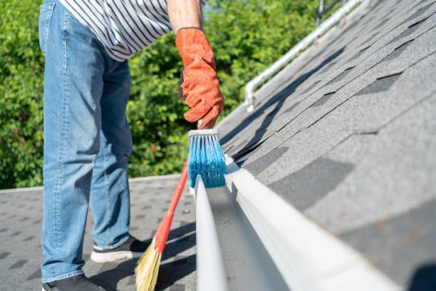 Person cleans roof gutter with a brush, wearing gloves.