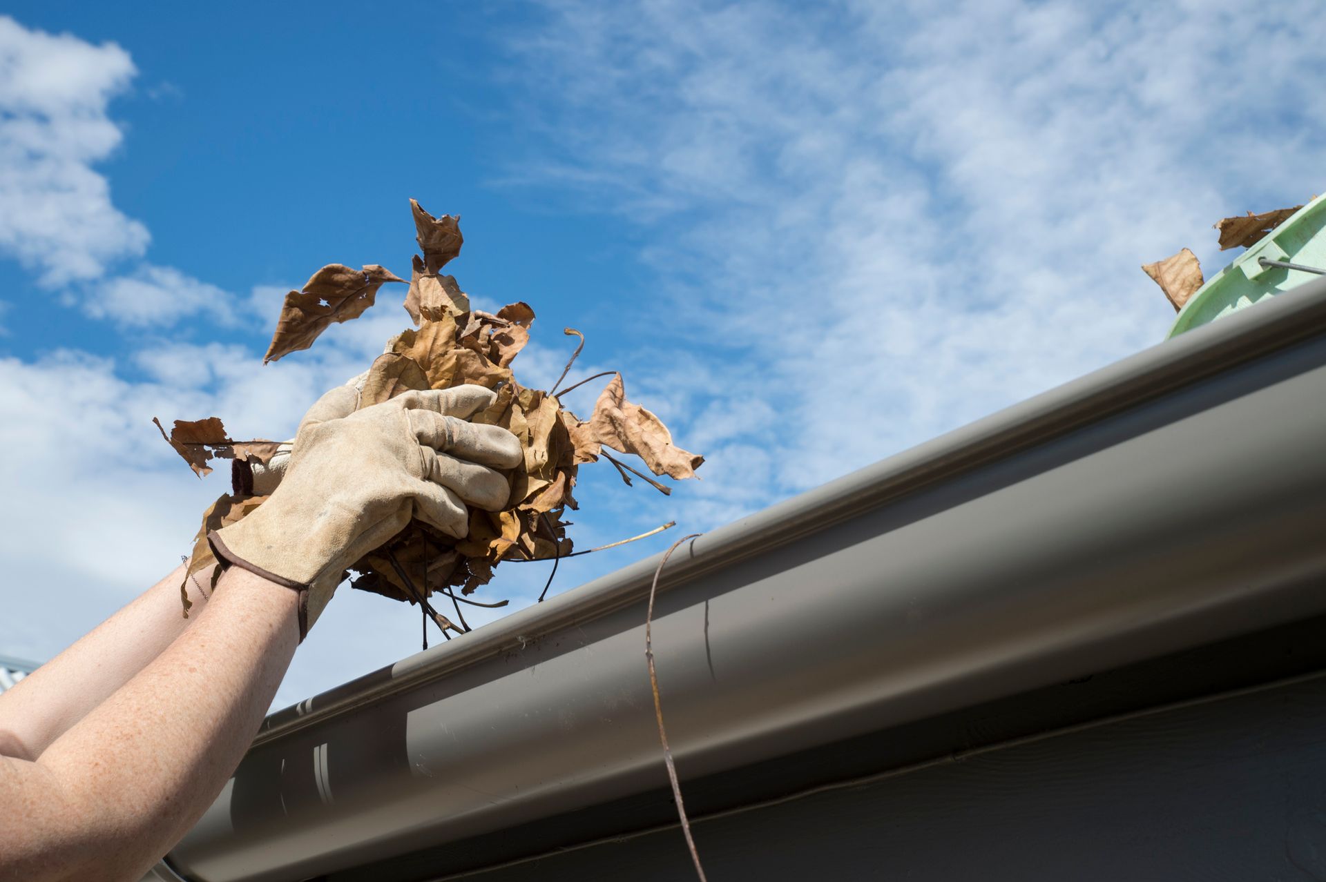 Person wearing gloves cleaning leaves from a gutter against a blue sky.