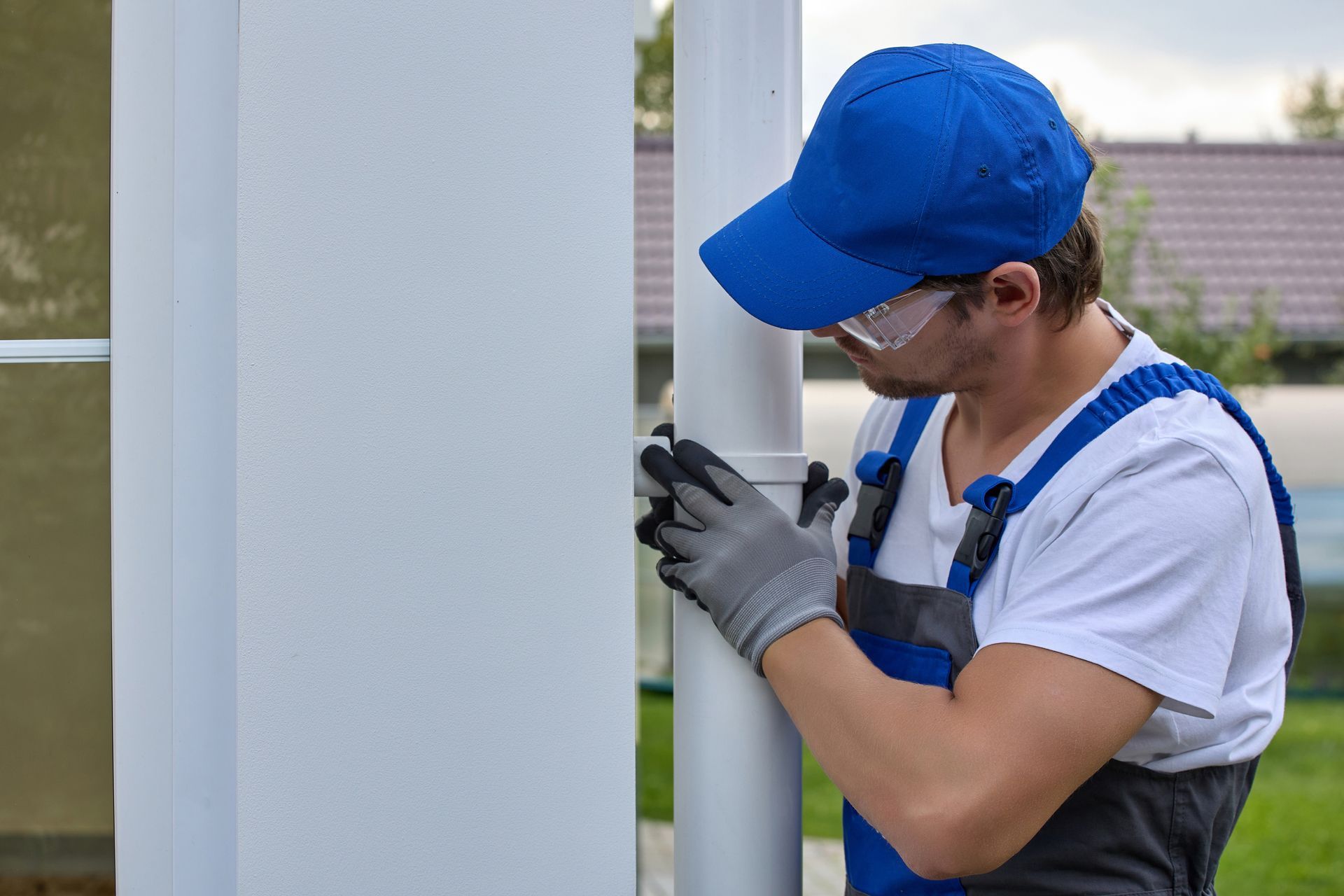 Man in blue cap and overalls installing a white gutter, outdoors.