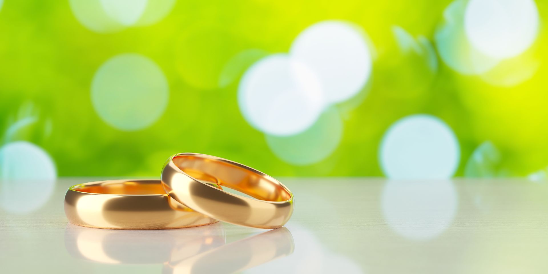 Two golden wedding rings on a reflective white surface with a blurred green background.