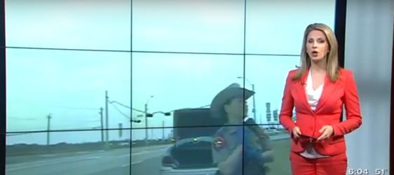 News anchor in red suit reporting in front of a video screen showing an outdoor scene.