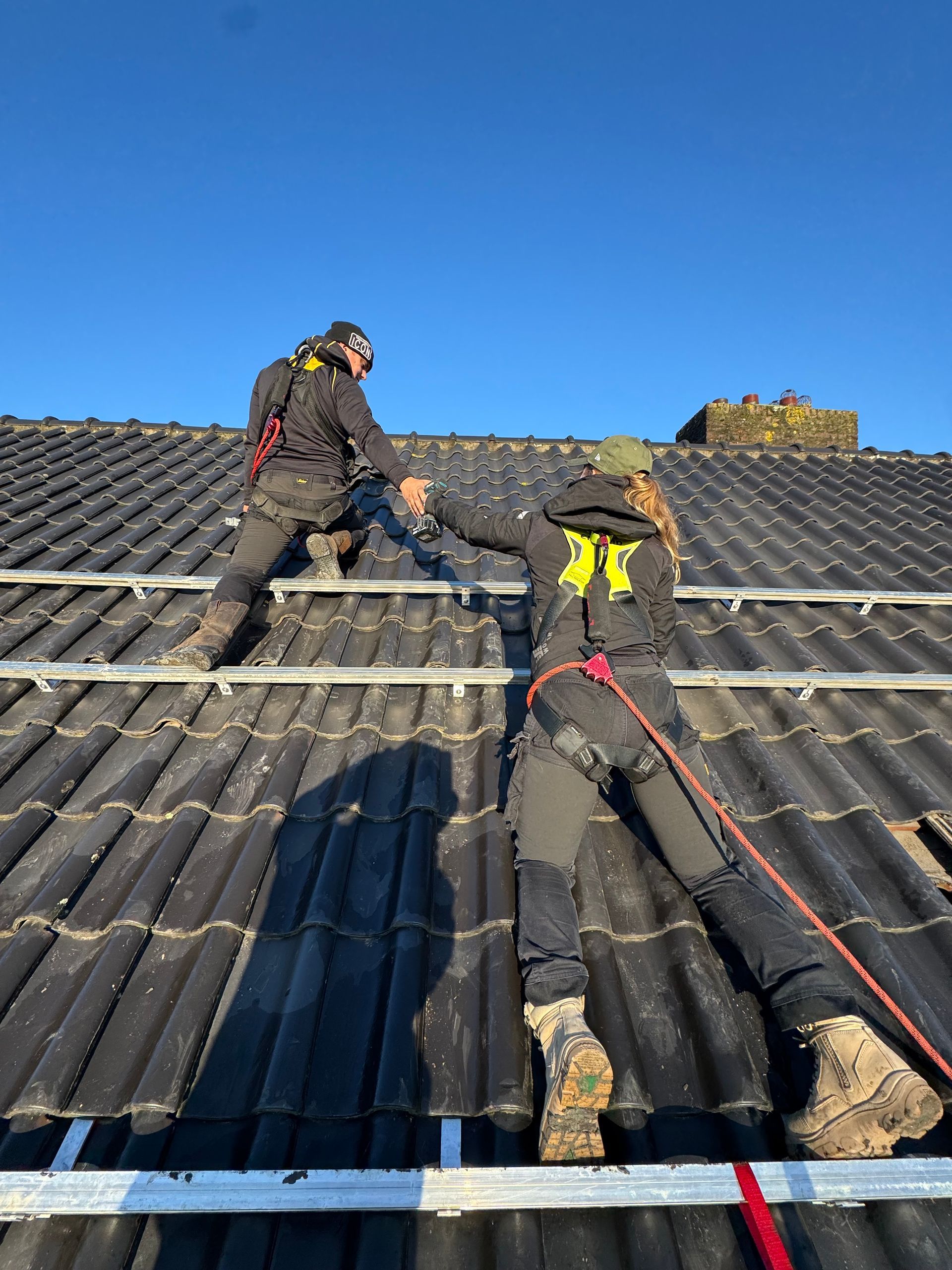Twee arbeiders, gezekerd met harnassen, installeren zonnepanelen op een dak op een zonnige dag.