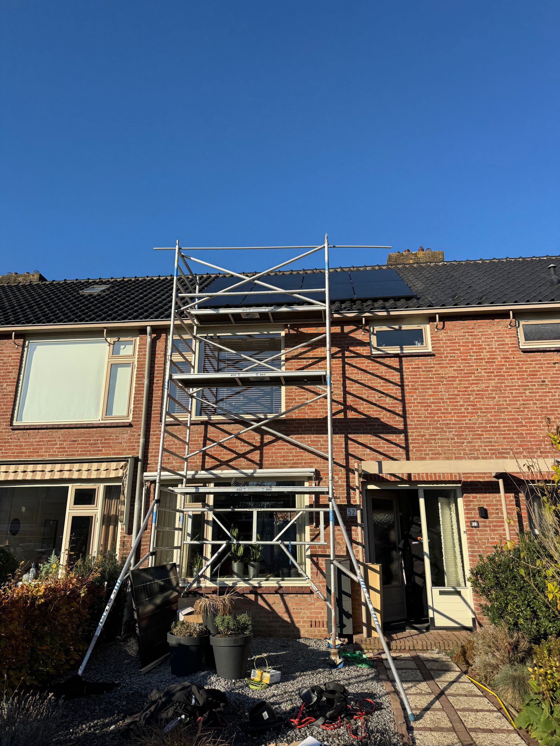 Een steiger tegen een bakstenen huis met beschadigde ramen en dak, onder een helderblauwe hemel.