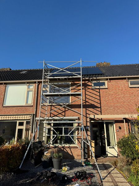 Bakstenen rijtjeshuis met steigers voor een raam; donker dak, zonnige dag.