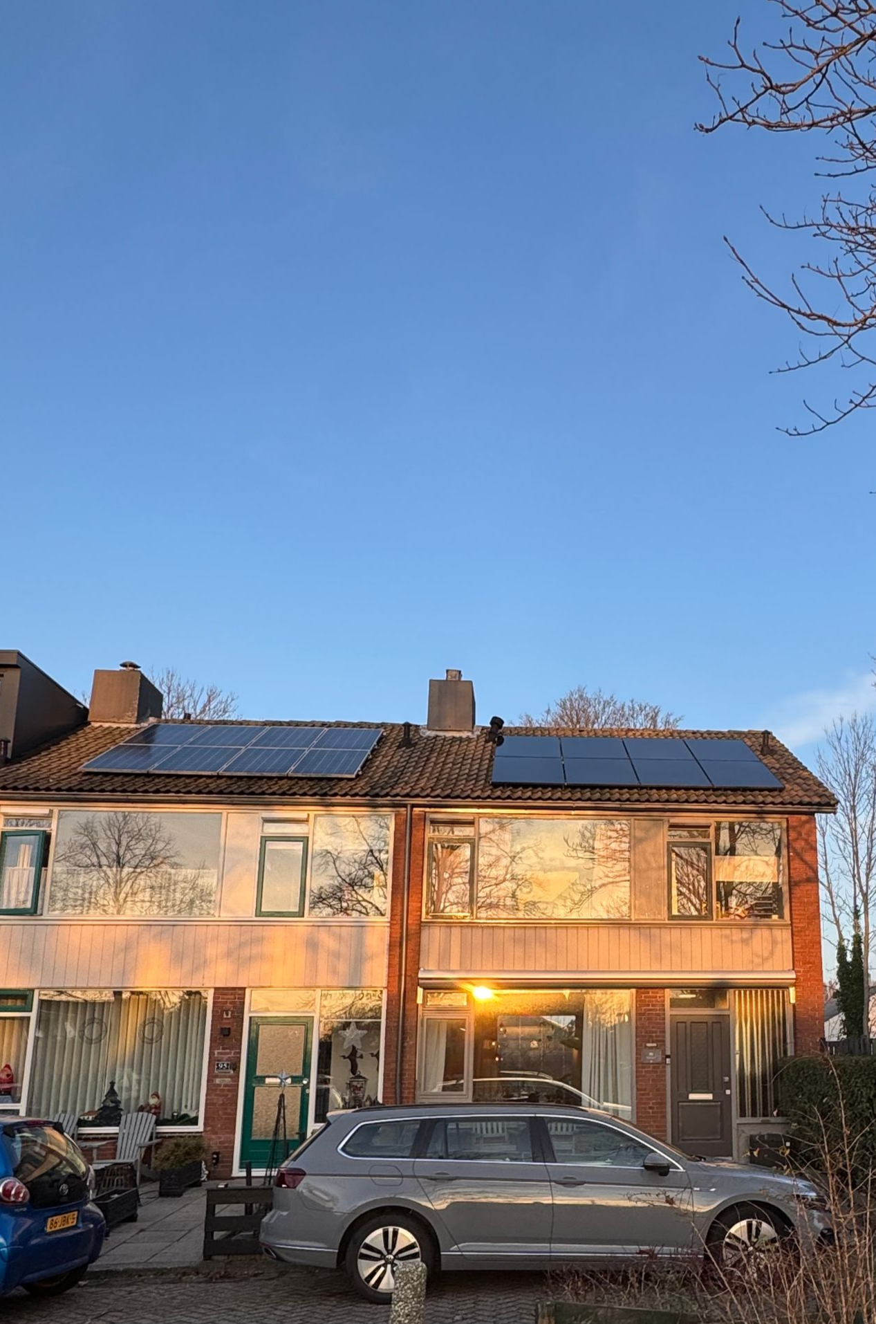 Rijtjeshuizen met zonnepanelen op het dak, badend in goudgeel zonlicht; er staat een auto voor geparkeerd.