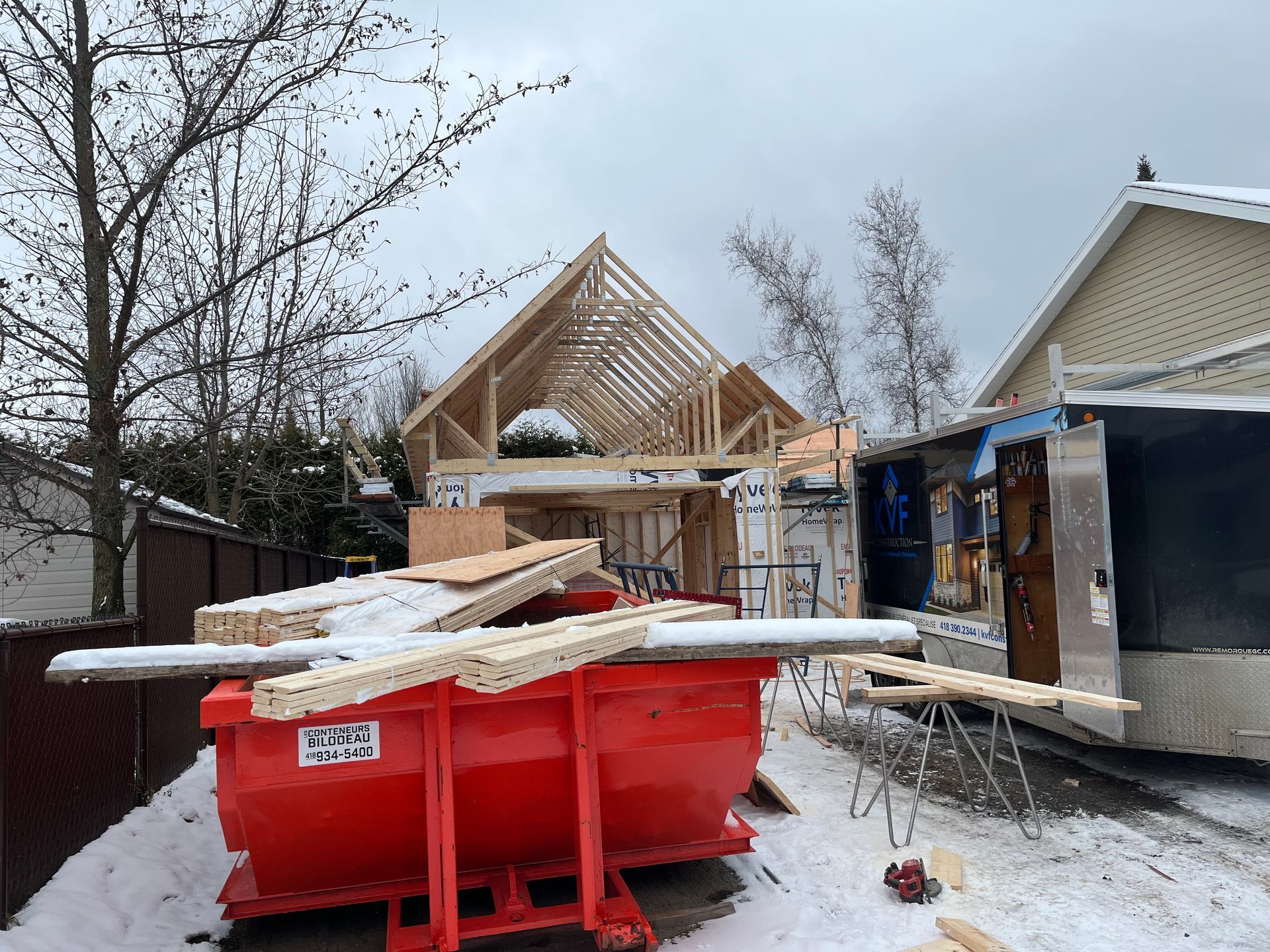 Une benne rouge est posée dans la neige devant une maison en construction.
