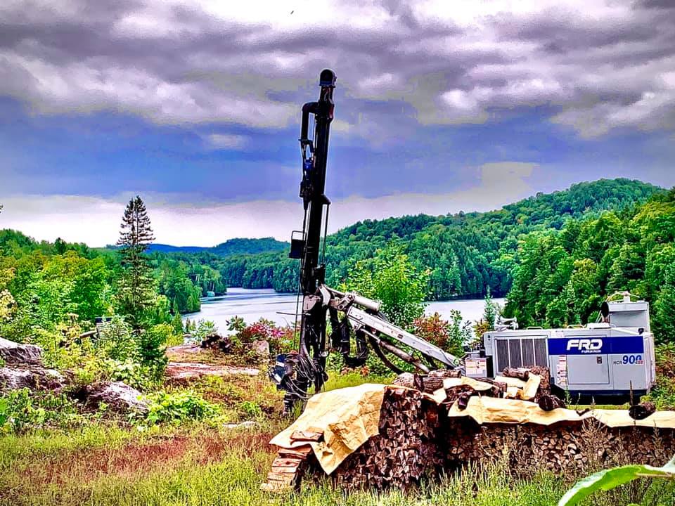 Plateforme de forage à flanc de colline surplombant un lac et des montagnes boisées sous un ciel nuageux.