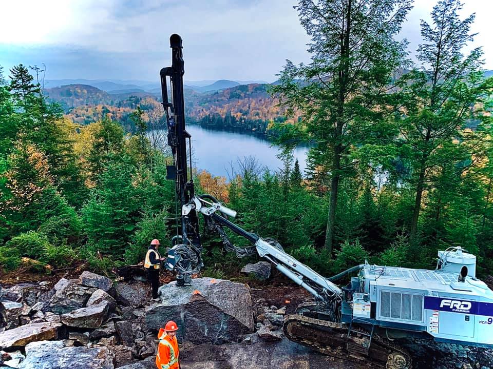 Machine de forage sur un flanc de colline rocailleux surplombant un lac, ouvriers en gilets orange. Forêt en arrière-plan.