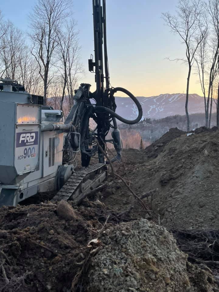 Une foreuse à flanc de colline, creusant le sol ; des montagnes en arrière-plan, ciel couvert.