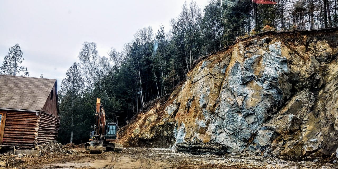 Chantier de construction avec une falaise rocheuse, des arbres et un bâtiment en bois sous un ciel nuageux. Une pelleteuse est présente.