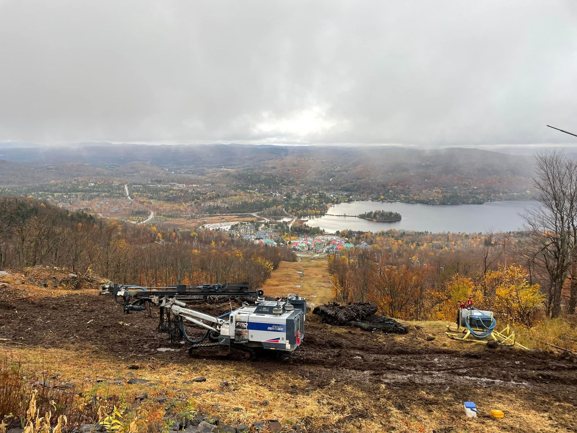 Engins de chantier à flanc de colline, surplombant une ville et un lac sous un ciel nuageux.
