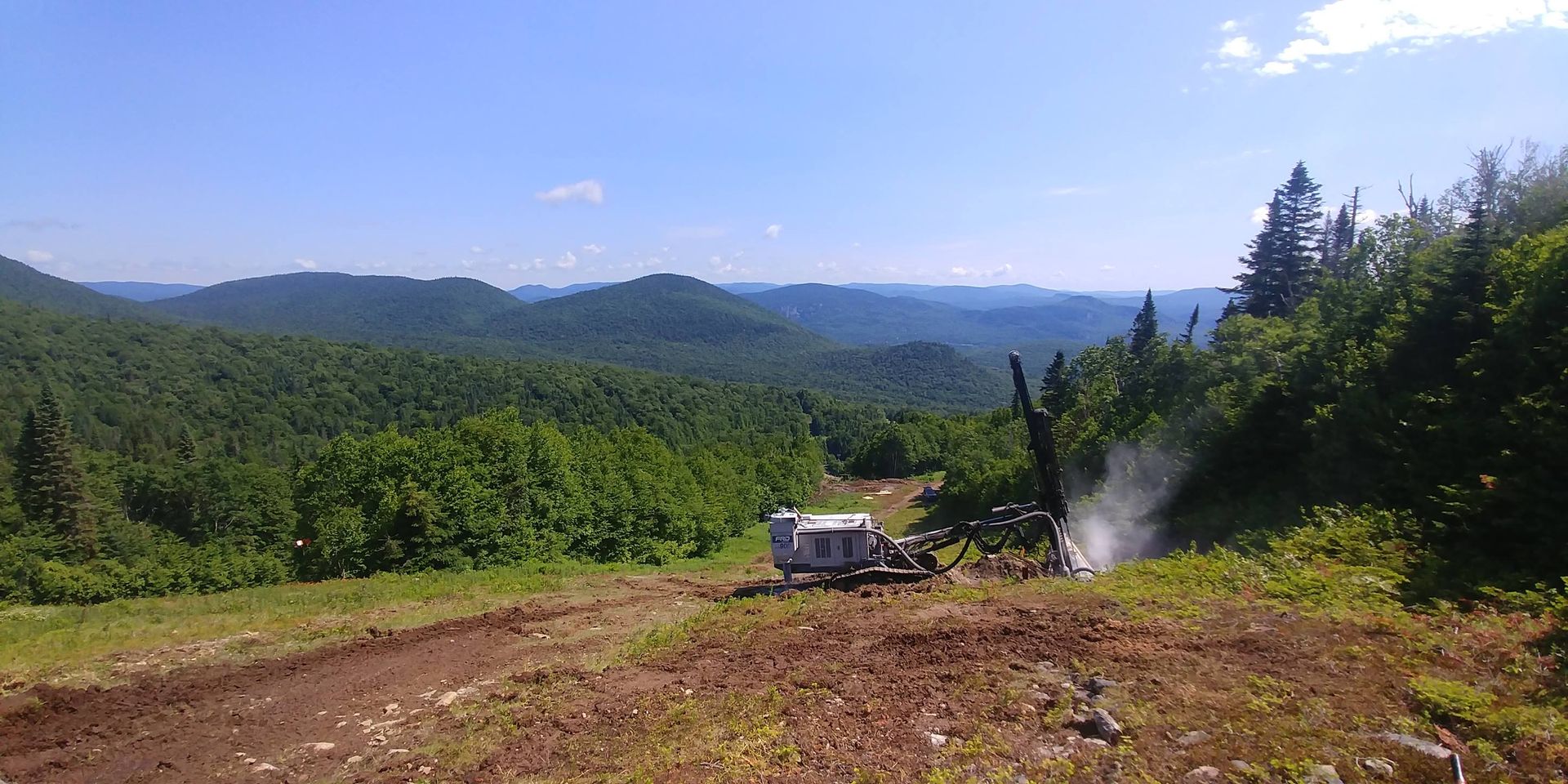 Un paysage montagneux avec des arbres verdoyants luxuriants et un ciel bleu, un sol de terre au premier plan et une machine arrosant.