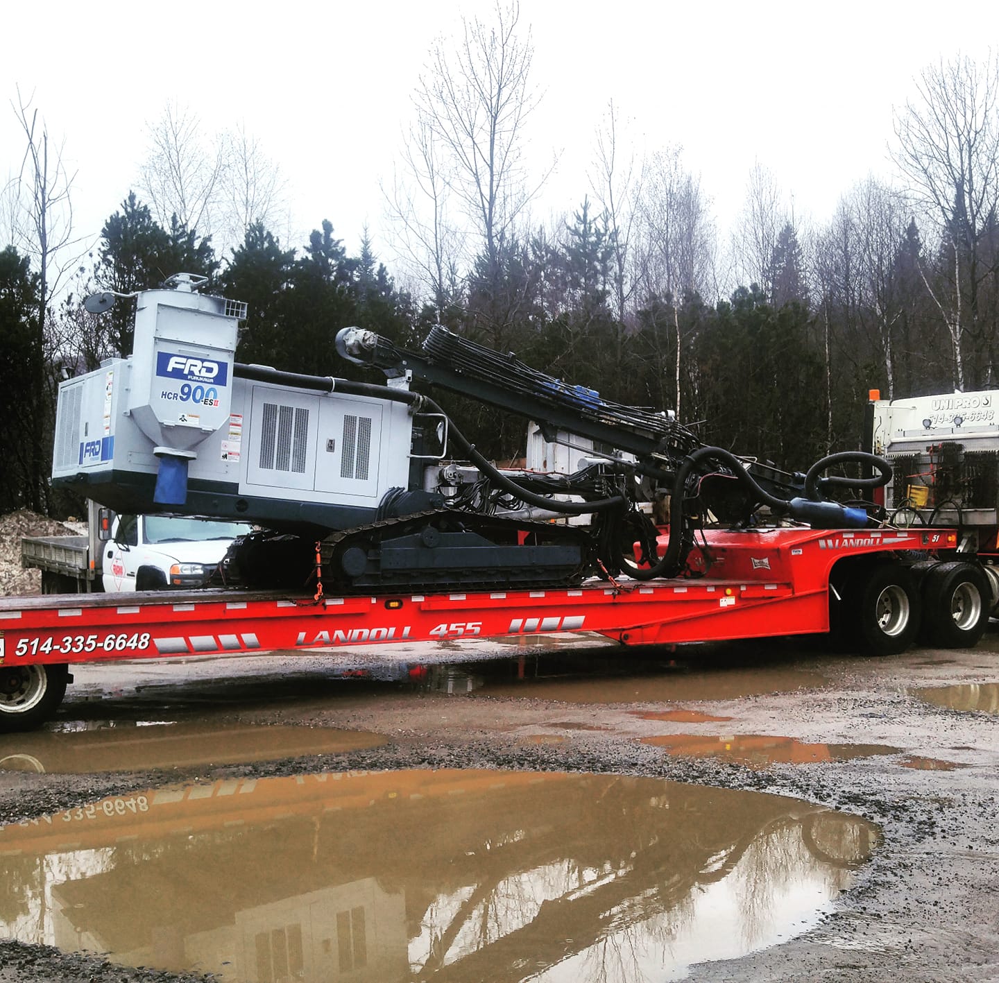 Une remorque plateau rouge transporte une foreuse dans une zone boueuse avec des arbres en arrière-plan.
