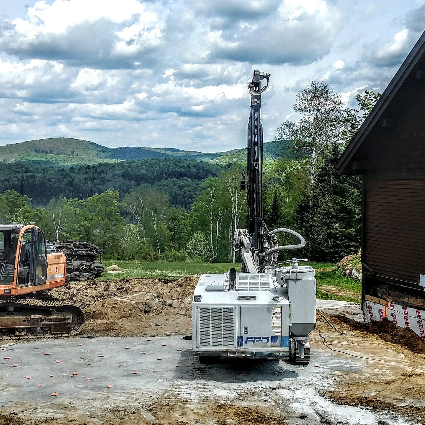 Engin de forage sur une dalle de béton près d'un bâtiment ; excavatrice à gauche, montagne boisée en arrière-plan.