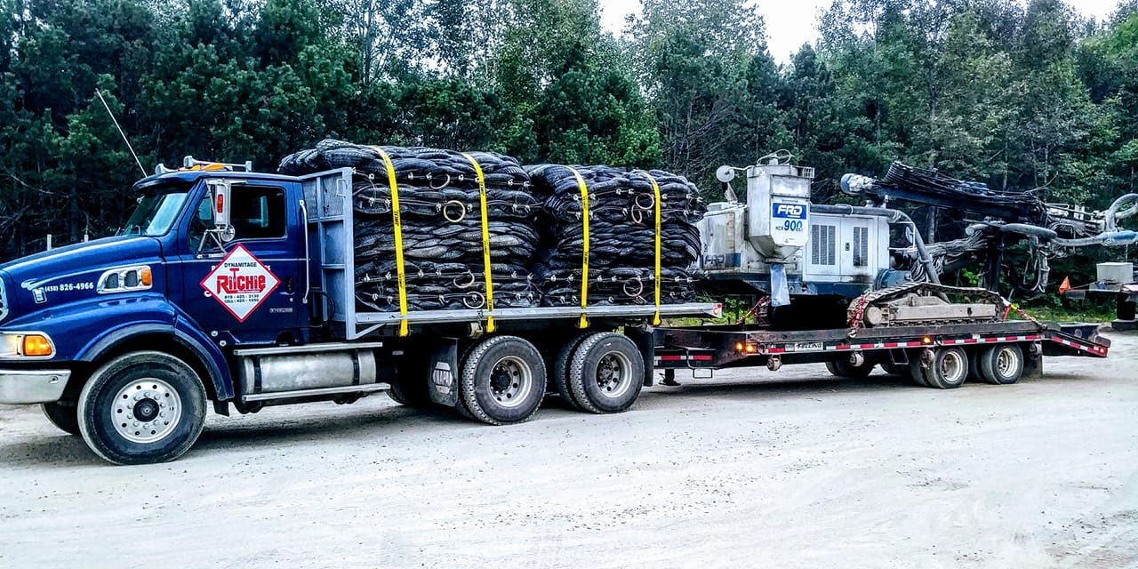 Un semi-remorque bleu chargé de tapis de caoutchouc noirs et de machines sur une remorque, stationné sur du gravier.