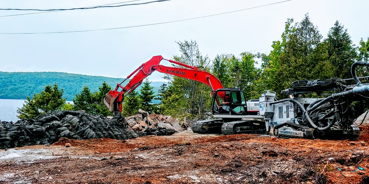Une excavatrice rouge et du matériel de forage sur un chantier de construction, avec des arbres et de l'eau en arrière-plan.