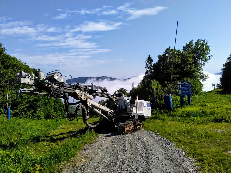 Une grosse foreuse blanche sur un chemin de gravier, avec une chaîne de montagnes masquée par les nuages ​​en arrière-plan.