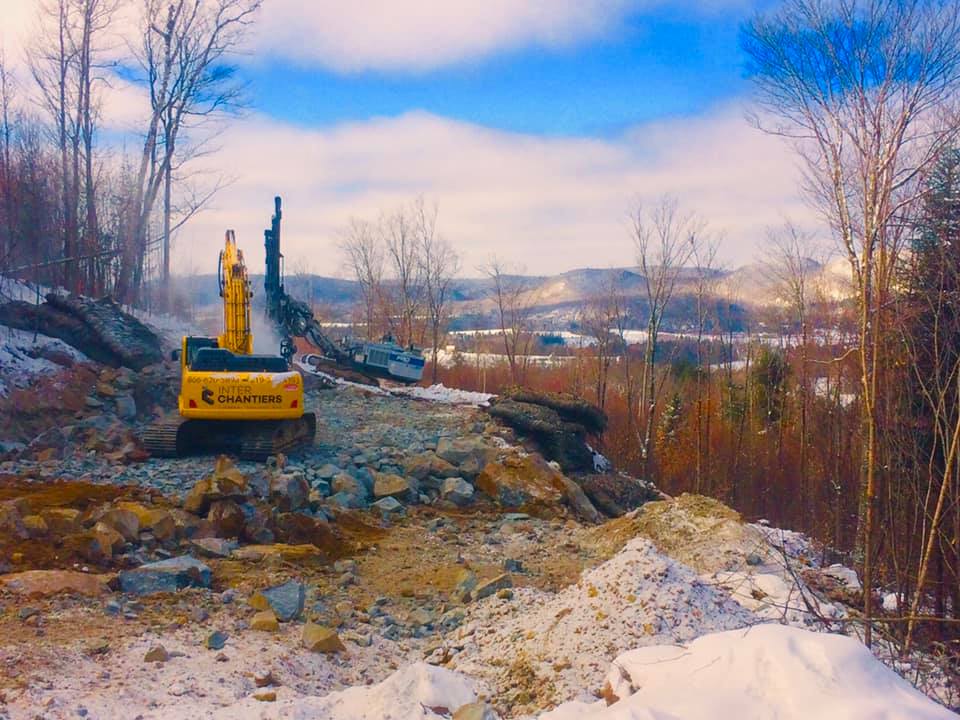 Une pelleteuse jaune casse des rochers sur un flanc de colline enneigé surplombant un village au loin.