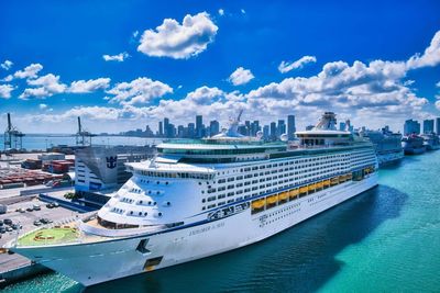 A large cruise ship is docked in a harbor with a city skyline in the background.