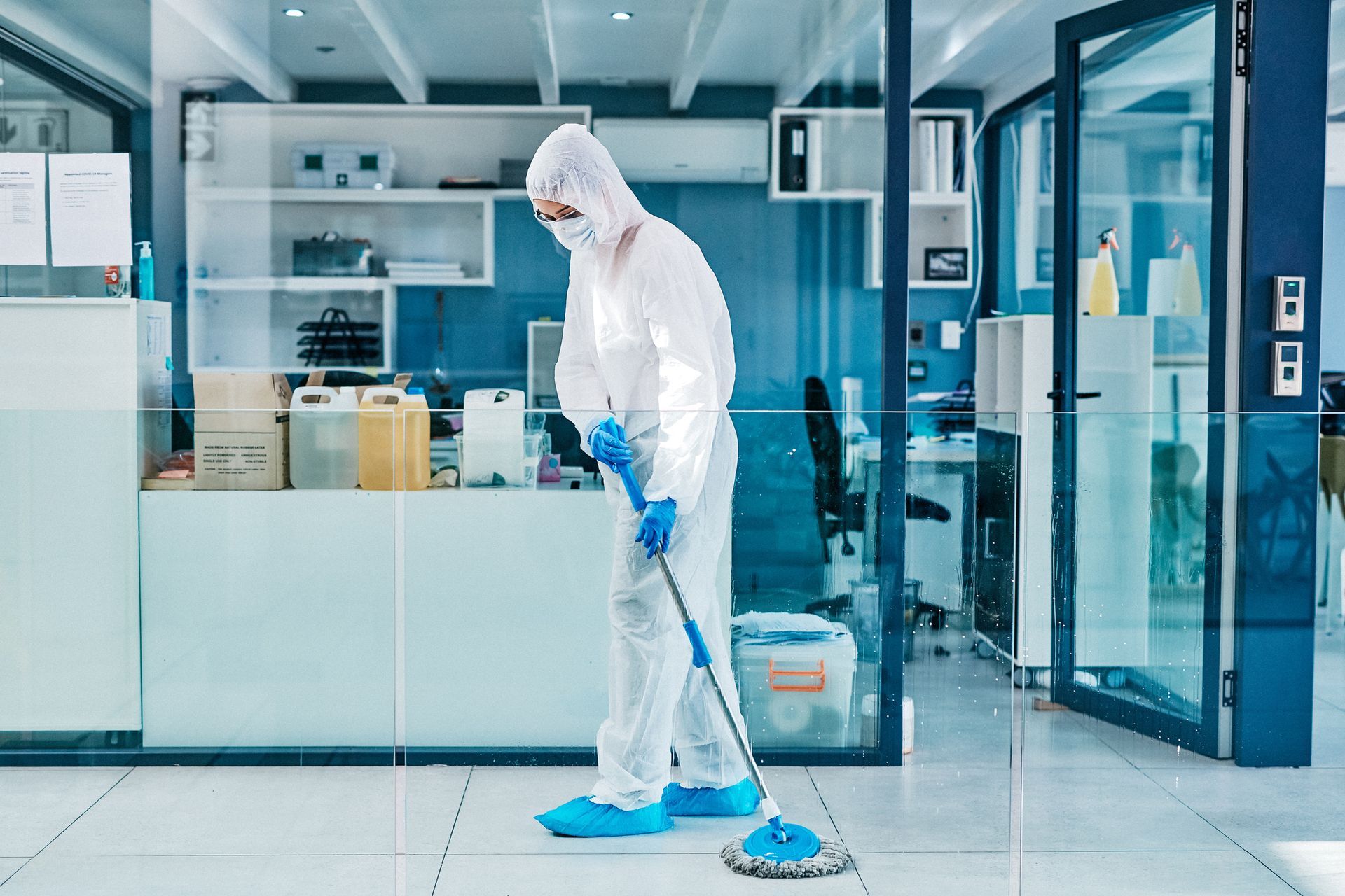 Person in hazmat suit mops an office floor. Bright, sterile environment; white, blue colors.