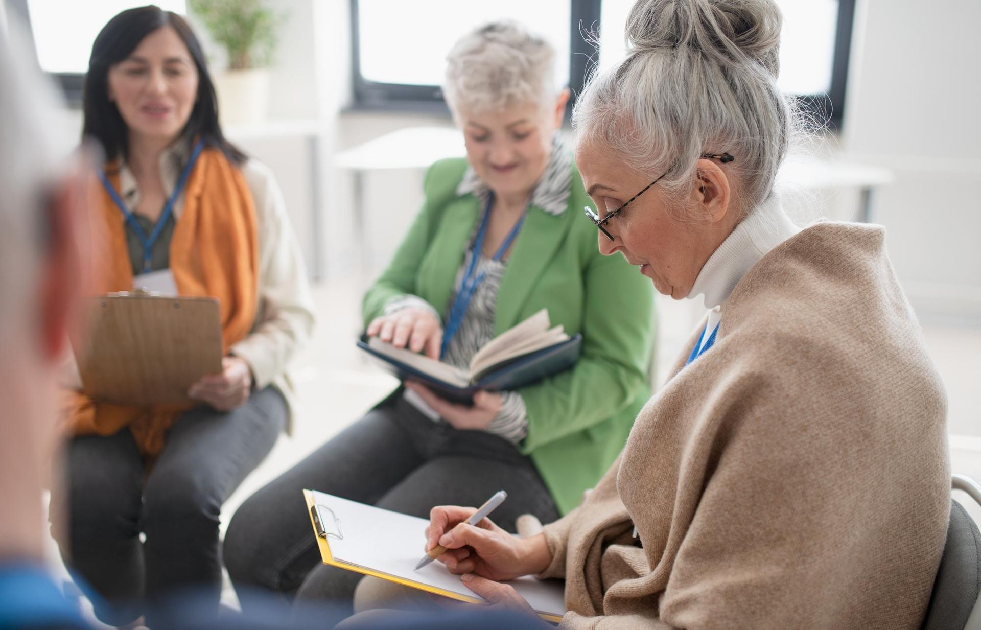 Group of older women sitting in a circle, one taking notes, another reading a book, smiling. Indoors setting.